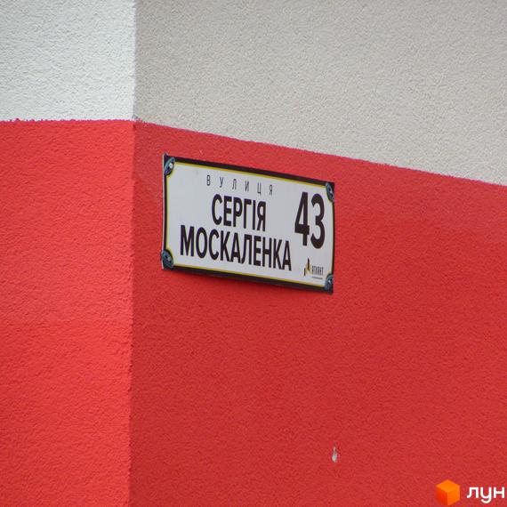 Вуличний знак з назвою вулиці Сергія Москаленка та номером будинку 43 на тлі червоної стіни. Напис вказує на розташування житлового комплексу.