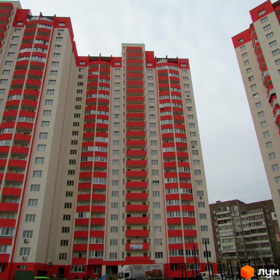 Високі будинки ЖК Олімпійський з червоними балконами на тлі сірого неба. На території комплексу видно зелень та припарковані автомобілі.