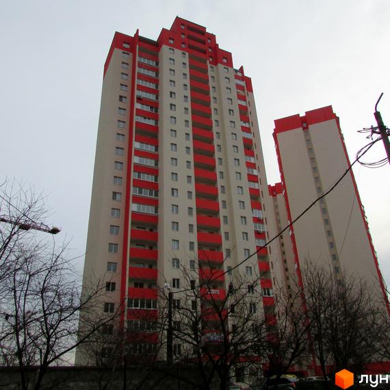Висотна будівля ЖК Олімпійський з червоними балконами та дахом. На передньому плані видно дерева, а на задньому - сіре небо.