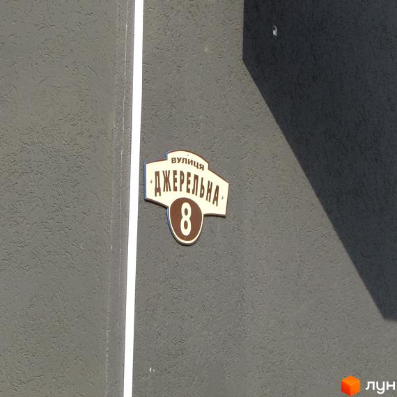 Вивіска з назвою вулиці та номером будинку на фасаді будівлі. Сірий фасад з білою смугою. На вивісці напис «вулиця Джерельна 8».