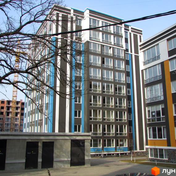 Житловий комплекс Фортуна-2 з фасадом у смужку. На передньому плані – гаражі та дерева, на задньому – будівництво.