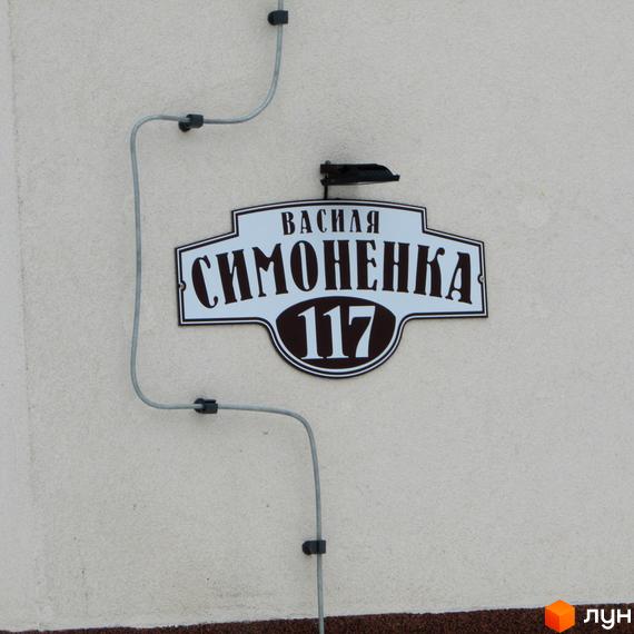 Вуличний знак з написом «Василя Симоненка 117» на фасаді будинку. Поруч прокладено кабель.