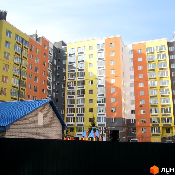 Багатоповерховий будинок з різнокольоровими фасадами. На території розташований дитячий майданчик та невелика будівля з синім дахом.