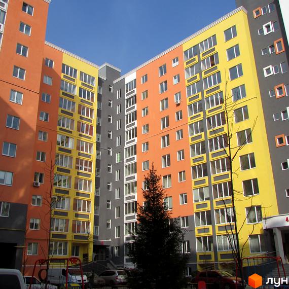 Яскравий житловий комплекс з різнокольоровими фасадами. На прибудинковій території є дерева, парковка та дитячий майданчик.
