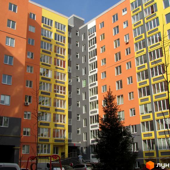 Яскравий багатоповерховий будинок житлового комплексу Синергія-2 з великою кількістю вікон. На прибудинковій території ростуть дерева та є парковка для автомобілів.