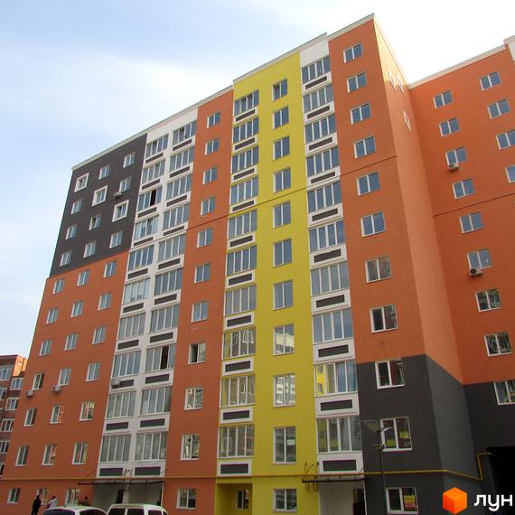 Багатоповерховий будинок житлового комплексу Синергія-2 з різнокольоровим фасадом та великою кількістю вікон. На території є парковка та зелені насадження.