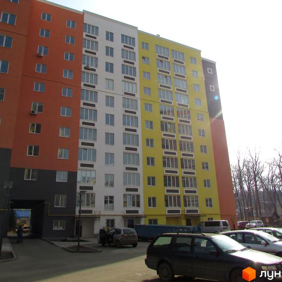 Багатоповерховий будинок з різнокольоровим фасадом. На прибудинковій території розташовані припарковані автомобілі та зелені насадження.