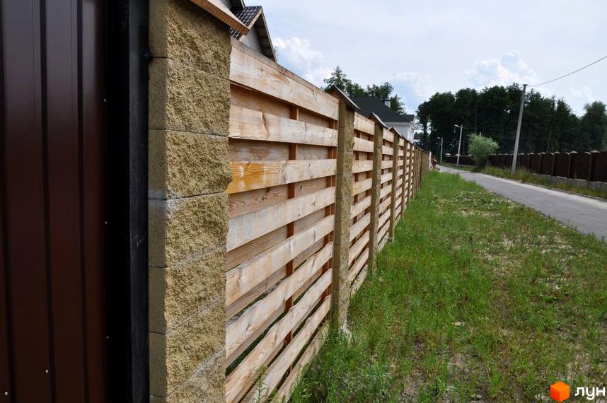 Огорожа з дерев'яних дощок та кам'яних стовпів. Поруч дорога та зелена трава.