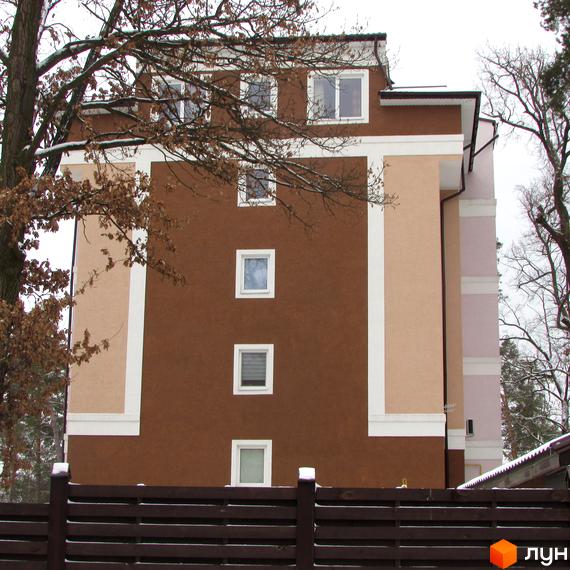 Житловий комплекс Київський дворик. Фасад будинку з коричневою та бежевою штукатуркою, білими декоративними елементами та квадратними вікнами. На передньому плані дерев'яний паркан.