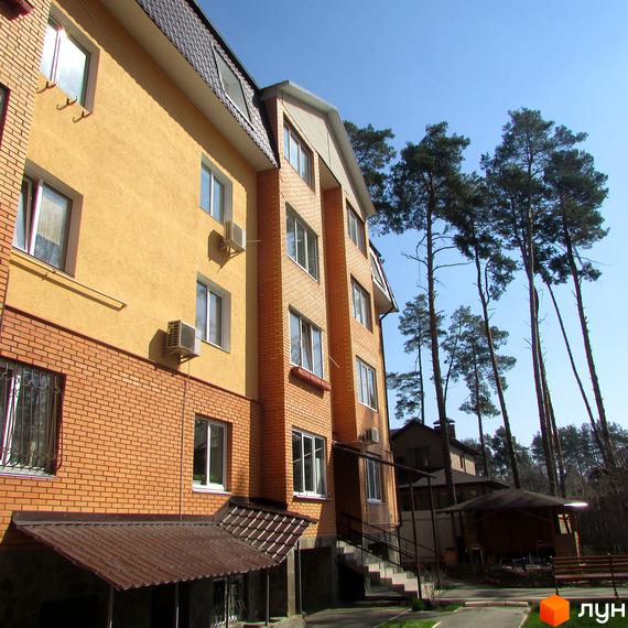 Фасад будинку з жовтою штукатуркою та цегляною обробкою, вікнами та кондиціонерами. Навколо будинку зелені дерева та блакитне небо. Прибудинкова територія з доріжками та навісом.