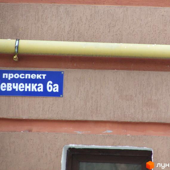 Фасад новобудови просп. Шевченка, 6а, з жовтою трубою над синьою табличкою з назвою вулиці. Будинок має коричневу штукатурку та темне вікно.