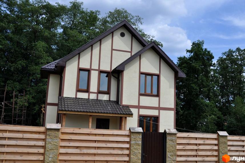 Двоповерховий будинок з коричневими вікнами та дахом. Перед будинком дерев'яний паркан з кам'яними стовпами, за ним зелені дерева.