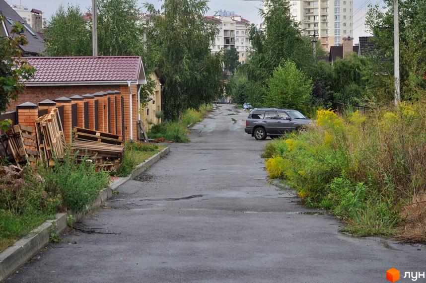 Вулиця з будинками та припаркованим автомобілем. На передньому плані видно цегляну будівлю з коричневим дахом та палетами.