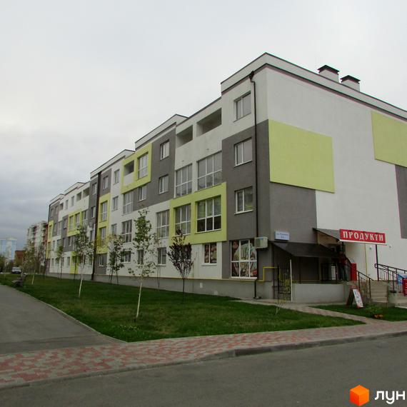 Зовнішній вигляд житлового комплексу Сан Сіті з фасадом, що поєднує сірий, білий та салатовий кольори. Перед будинком розташована зелена зона з деревами та доріжка з бруківки.