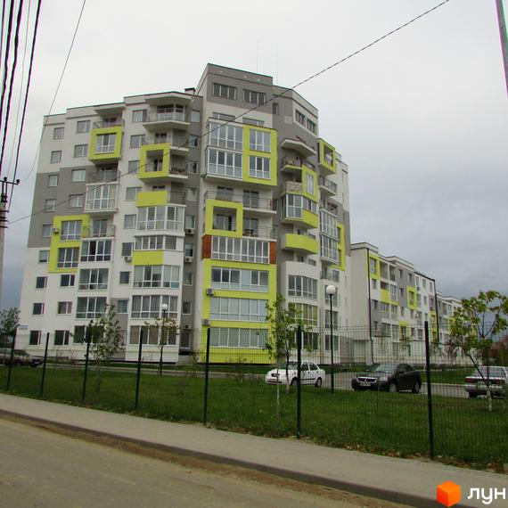 Житловий комплекс Сан Сіті: багатоповерховий будинок з яскраво-жовтими елементами фасаду та скляними балконами. Навколо будинку зелена зона з деревами та парканом, видно припарковані автомобілі.