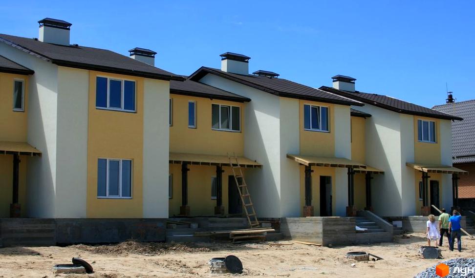 Зображення житлового комплексу з двоповерховими будинками з жовтими та білими стінами, коричневими дахами та трубами. На передньому плані — піщана територія та люди.