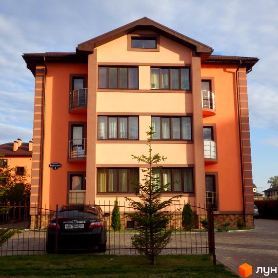 Зовнішній вигляд будинку житлового комплексу Волошковий з оранжевим фасадом та коричневими вікнами. На прибудинковій території видно паркан, дерево та автомобіль.