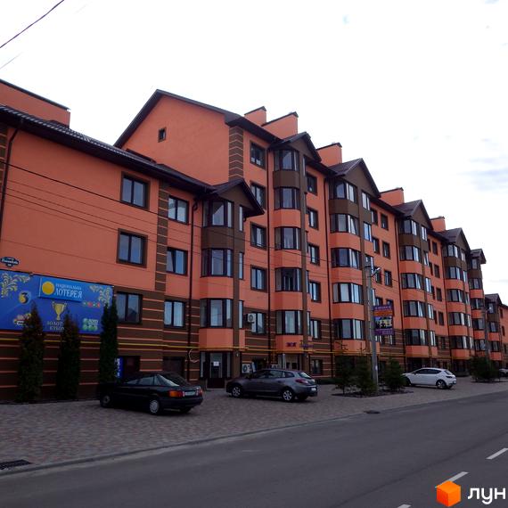 Житловий комплекс Волошковий - це багатоповерховий будинок з помаранчевим фасадом та коричневими елементами. Перед будинком розташована парковка та зелені насадження.