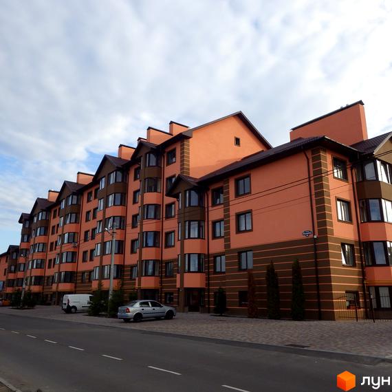 Зовнішній вигляд житлового комплексу Волошковий з фасадом персикового кольору та коричневими елементами. Навколо новобудови розташована прибудинкова територія з дорогою та автомобілями.