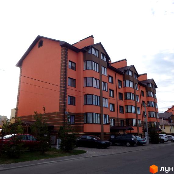 Житловий комплекс Волошковий – це багатоповерхова новобудова з оранжевим фасадом та коричневими елементами. Перед будинком розташована прибудинкова територія з деревами та припаркованими автомобілями.