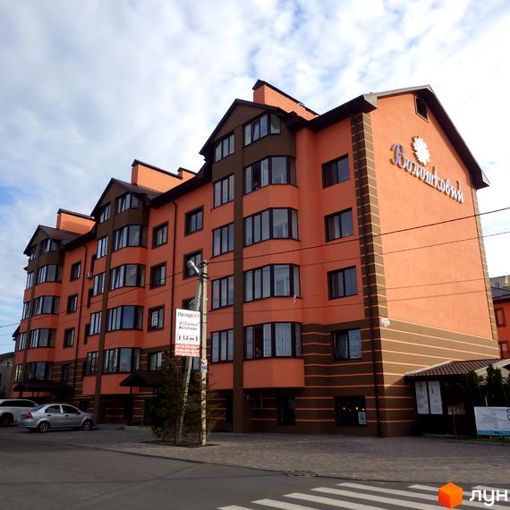 Зовнішній вигляд житлового комплексу Волошковий з оранжевим фасадом та коричневими елементами. Навколо будівлі розташована прибудинкова територія з дорогою та пішохідним переходом.