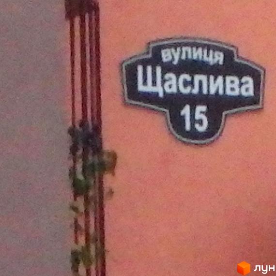 Назва вулиці Щаслива, будинок 15. На фасаді будинку видно табличку з назвою вулиці та номером будинку. Поруч з табличкою ростуть рослини.