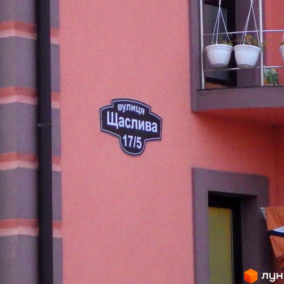 На фасаді будинку табличка з назвою вулиці Щаслива та номером будинку 17/5. На балконі висять білі горщики з квітами.