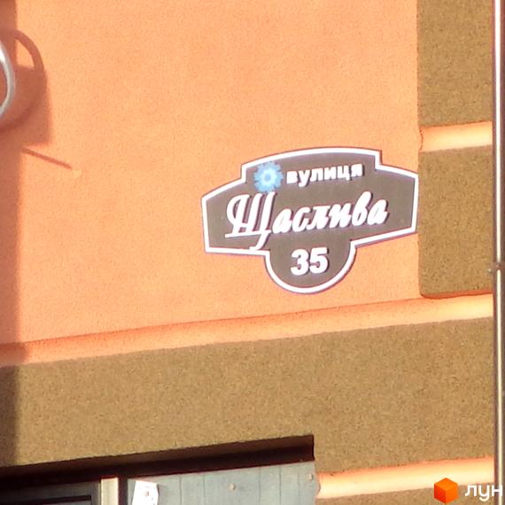Назва вулиці Щаслива, 35 на фасаді будинку. Новобудова з оранжевим фасадом та зеленим оздобленням.
