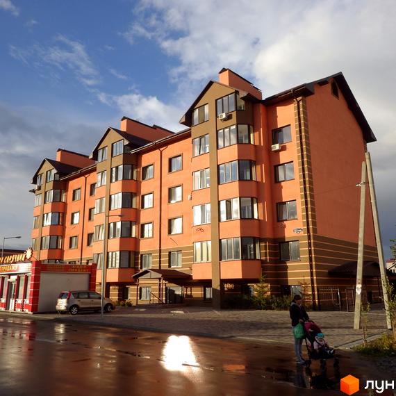 Житловий комплекс Волошковий - це яскрава новобудова з помаранчевим фасадом та коричневими елементами. На фото видно частину прибудинкової території з дорогою та дитиною з візочком.