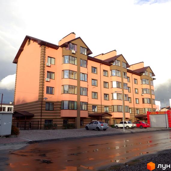 Зовнішній вигляд трисекційного житлового комплексу Волошковий з фасадом персикового кольору та коричневими акцентами. Перед будинком розташована прибудинкова територія з паркуванням.