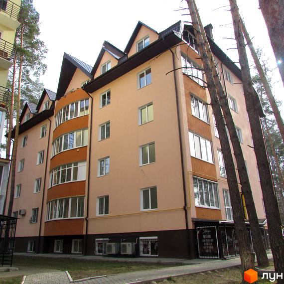 Зовнішній вигляд житлового комплексу Адміральський з фасадом персикового кольору та коричневим дахом. Навколо будинку ростуть високі сосни, а біля входу розташована прибудинкова територія з доріжками.