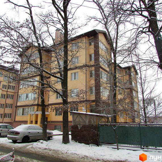 Фасад Житлового комплексу Високий взимку: світла будівля з темними декоративними елементами, на передньому плані засніжена територія з деревами та авто.