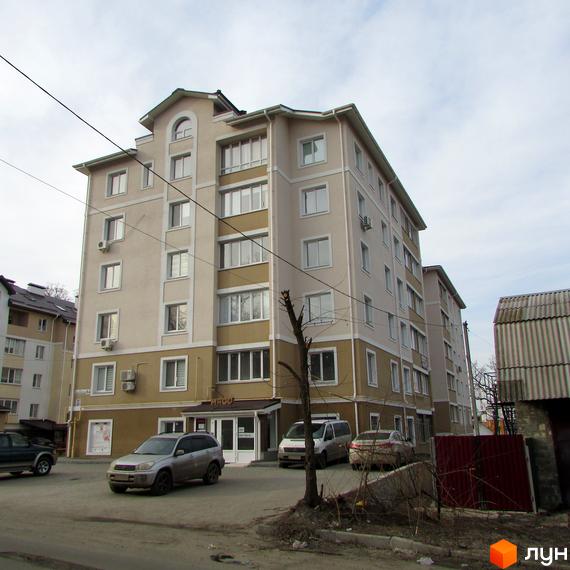 Багатоповерховий житловий будинок світлого кольору з коричневими акцентами та численними вікнами. Біля будинку видно припарковані автомобілі та голе дерево.