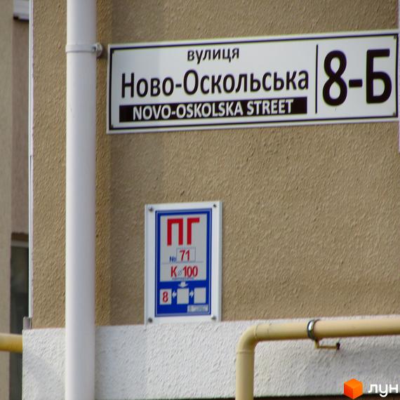 Частина фасаду житлового комплексу на вул. Новооскольська, 8б з табличкою вулиці. На стіні також розміщені технічні позначки та труби.