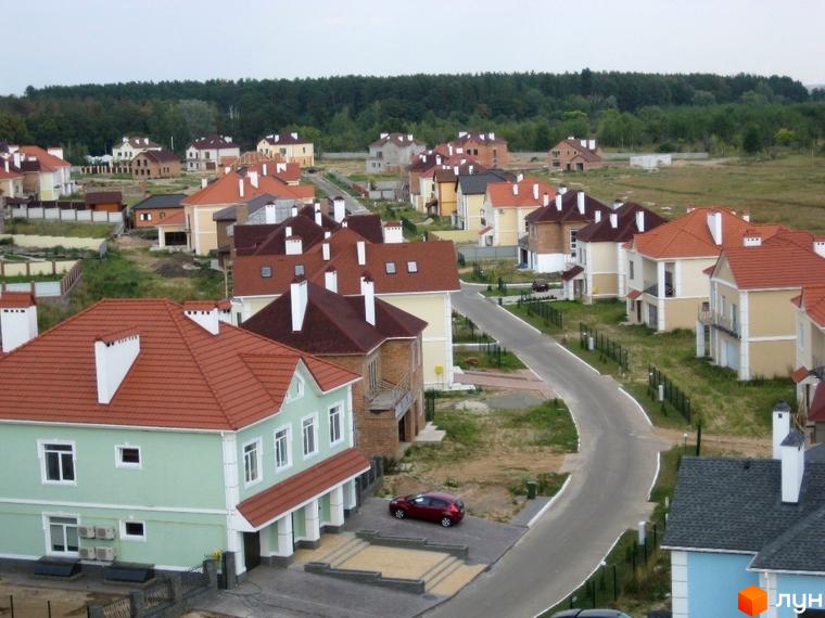 Котеджне містечко Grand Villas. Двоповерхові будинки з теракотовими дахами та білими димарями розташовані вздовж асфальтованої дороги. Навколо будівель зелені газони та дерева.