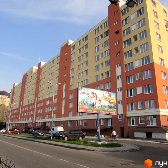 Зовнішній вигляд Житлового комплексу Вишневий затишок: багатоповерховий будинок з помаранчевим та жовтим фасадом, велика рекламна конструкція та зелена прибудинкова територія.