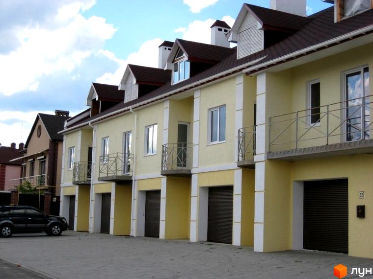 Зображення сторінки житлового комплексу КМ Гранд Віллас. Ряд будинків з гаражами та балконами, світлі фасади з темними дахами. Прибудинкова територія викладена плиткою.