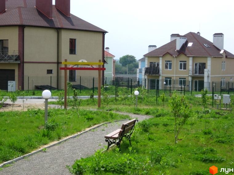 Двоповерховий будинок з коричневим дахом та світлими стінами. На прибудинковій території розташовані доріжки, лавки, зелені насадження та ліхтарі.