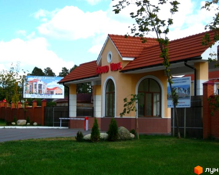 Вхідна група Містечко котеджів Grand Villas з червоним дахом та великими вікнами. На території зелена трава, дерева та рекламні банери.