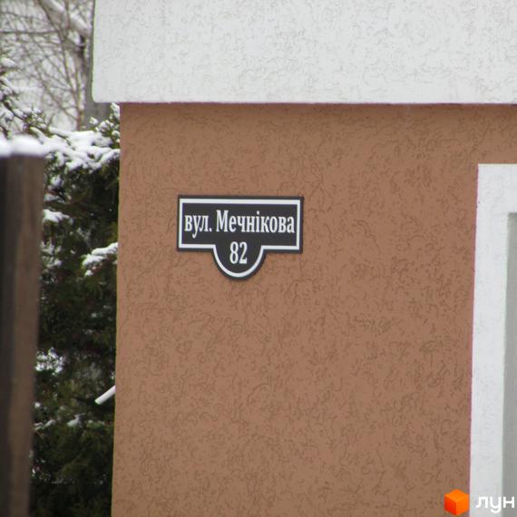 Житловий комплекс Ирпенский дворик: табличка з номером будинку 82 на вулиці Мечнікова на коричневій стіні. Навколо видно засніжені дерева.