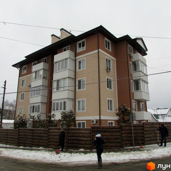 Житловий комплекс Ирпенский дворик: багатоповерховий будинок з коричневим дахом та бежево-коричневим фасадом. Перед ним дерев'яний паркан і засніжена територія з людьми.