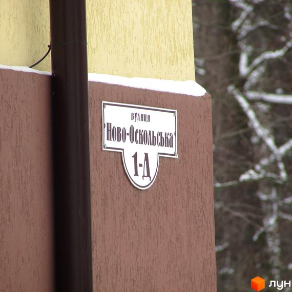 Табличка з адресою Ново-Оскольська 1-А на фасаді будинку Новобудови НовоОскольский. Стіна будівлі двоколірна, на задньому плані засніжені дерева.
