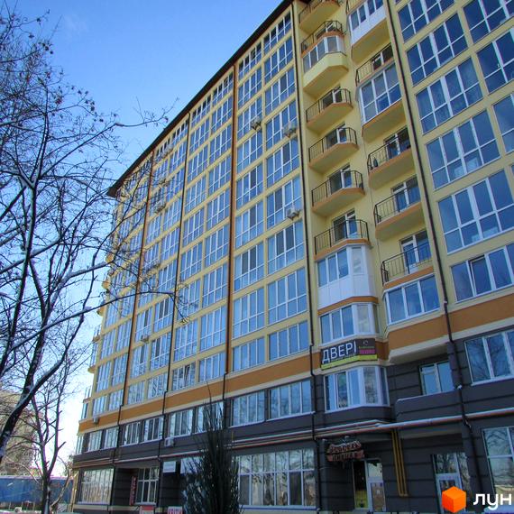 Фасад Новобудови Французький квартал з жовтими стінами, великими вікнами та балконами. На першому поверсі комерційні приміщення, поруч дерева.