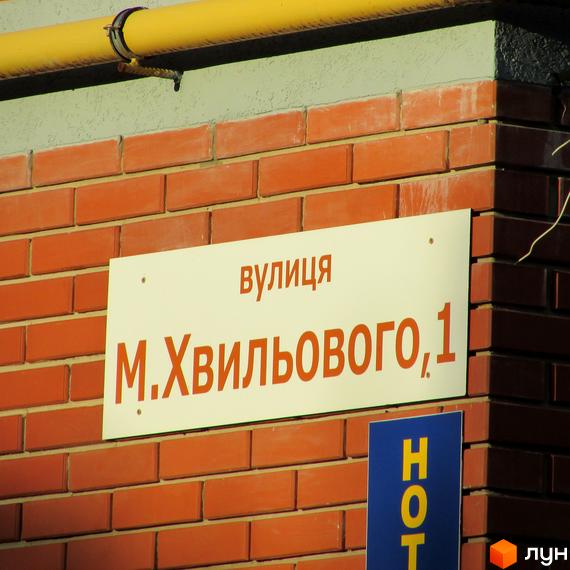 Вказівник вулиці Миколи Хвильового, 1, на цегляній стіні. Напис українською мовою на білій табличці. Поруч видно частину синьої таблички з написом 