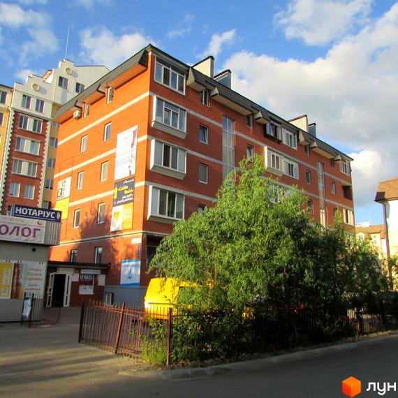 Зовнішній вигляд цегляного будинку з численними вікнами та мансардними поверхами. На прибудинковій території видно зелені насадження та паркан.