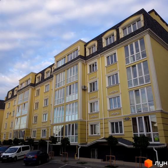 Фасад жовтого житлового комплексу Софіївський пасаж з великими вікнами та мансардними вікнами під сірим небом. На передньому плані видно паркувальні місця та дерева.