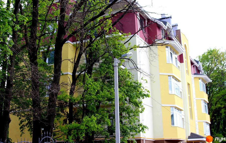 Зображення фасаду багатоповерхового будинку. Житловий комплекс Ваша квартира, з яскравими кольорами та великими вікнами. На передньому плані дерева.