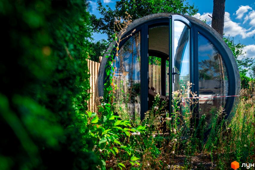 Кругла скляна споруда VISION розташована серед густої зелені та високої трави. Видно дерев'яний паркан та дерева на задньому плані.