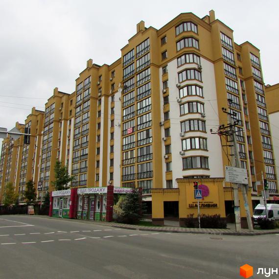 Житловий комплекс Щасливий на Петропавлівці. Фасад будинку в жовто-білих тонах, багато вікон та балконів. Поруч дорога та зелені насадження.