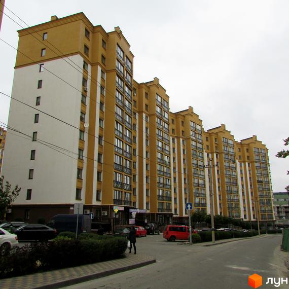 Зовнішній вигляд жовто-білого багатоквартирного будинку ЖК Щасливий на Петропавлівці. Прибудинкова територія з дорогою та зеленими насадженнями.